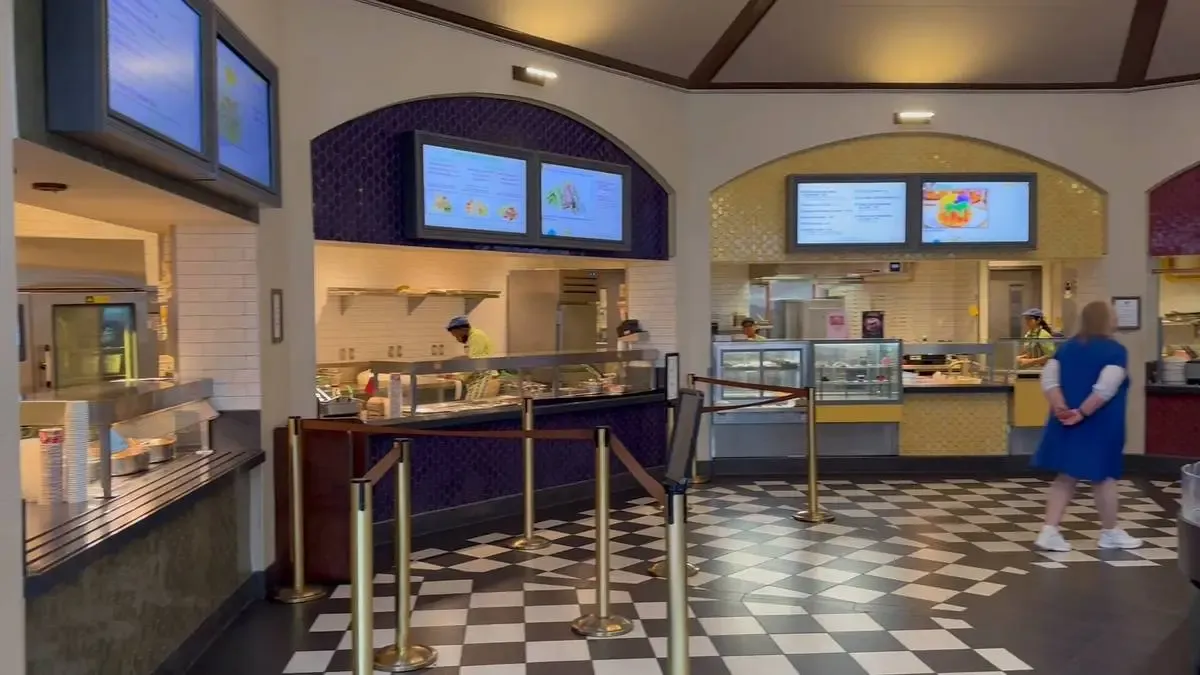 A cafeteria with a checkered floor features multiple food stations with digital menu screens above. A person in a blue dress stands nearby, while staff members work behind the counters preparing food.