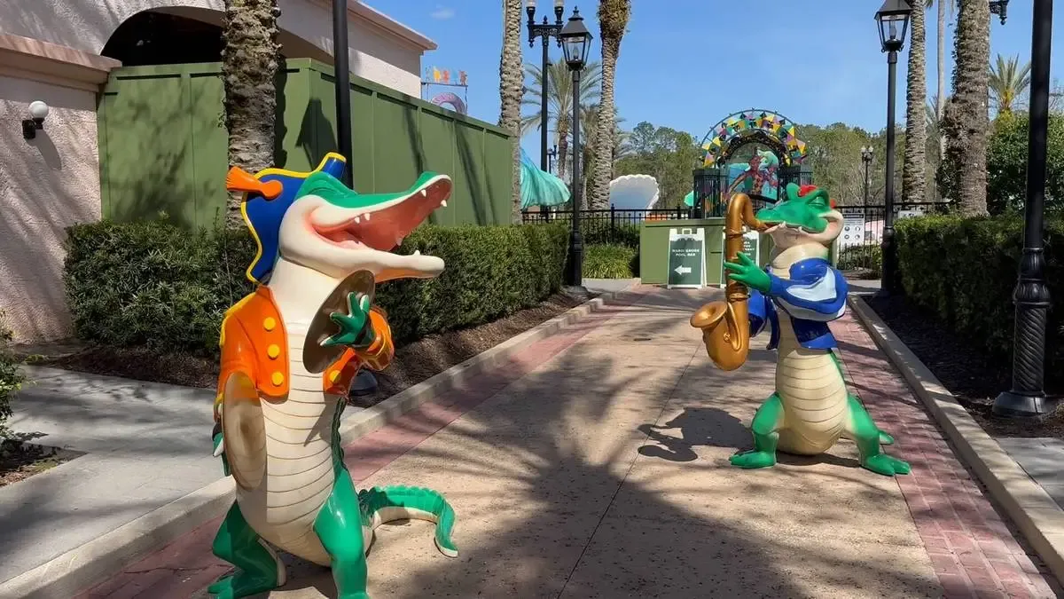 Two colorful alligator statues, one playing cymbals and the other a saxophone, stand on a sunny pathway lined with palm trees and lush greenery in a playful outdoor setting.