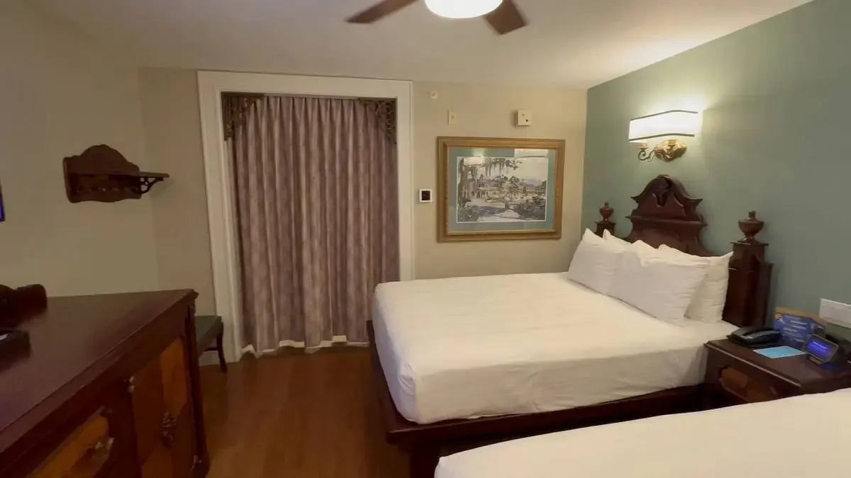 An hotel room featuring two double beds with ornate wooden headboards. A framed painting hangs on the wall above a bedside table with a phone and notepad. 