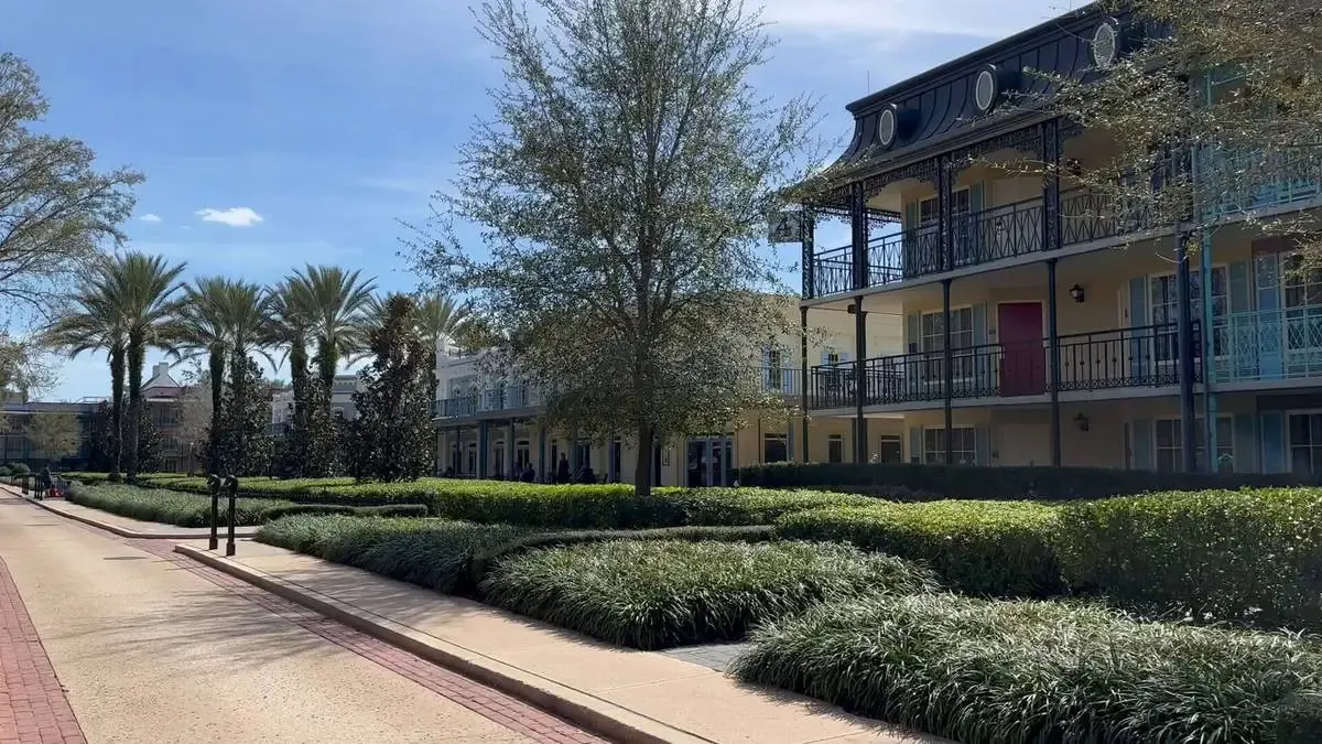 A walkway lined with palm trees leads to a building with ornate balconies and a red door, surrounded by lush greenery under a clear blue sky.