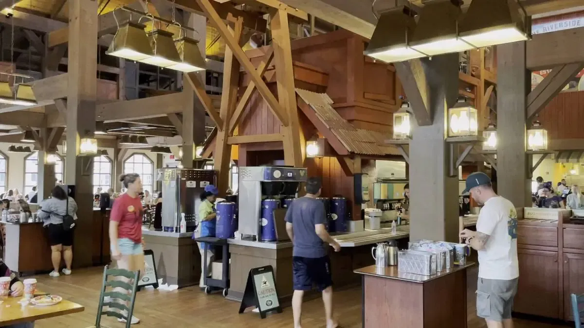 A busy cafeteria features several people moving around a beverage station with large coffee dispensers. The interior is rustic with wooden beams and hanging lanterns, creating a cozy atmosphere.