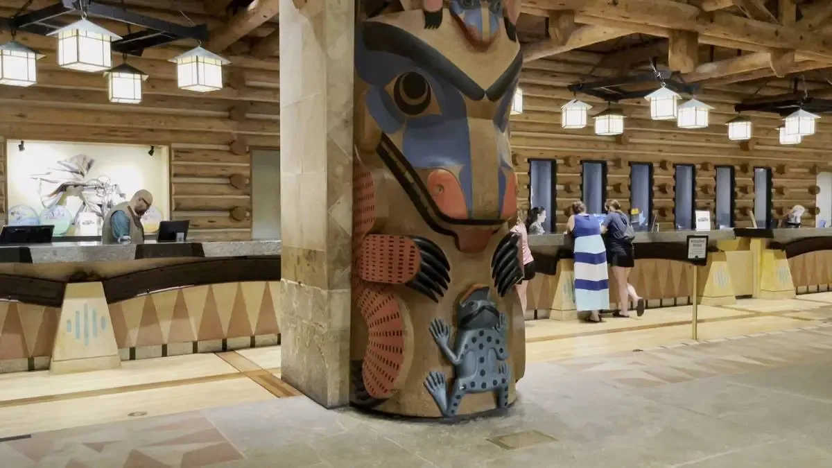 A hotel lobby features a large and colorful totem with intricate designs, standing by a rustic wooden reception desk. Several guests are interacting with staff behind the desk.