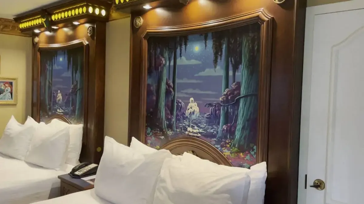 An hotel room featuring two beds with white pillows and sheets. Above each bed, there's a detailed, framed mural depicting a whimsical nighttime forest scene with a glowing waterfall.
