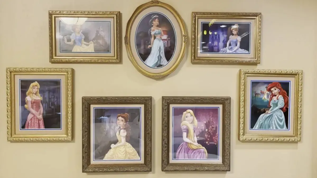 A collection of framed portraits featuring animated princesses arranged on a beige wall. The frames, in various ornate styles, include images of princesses in elegant poses and colorful gowns.