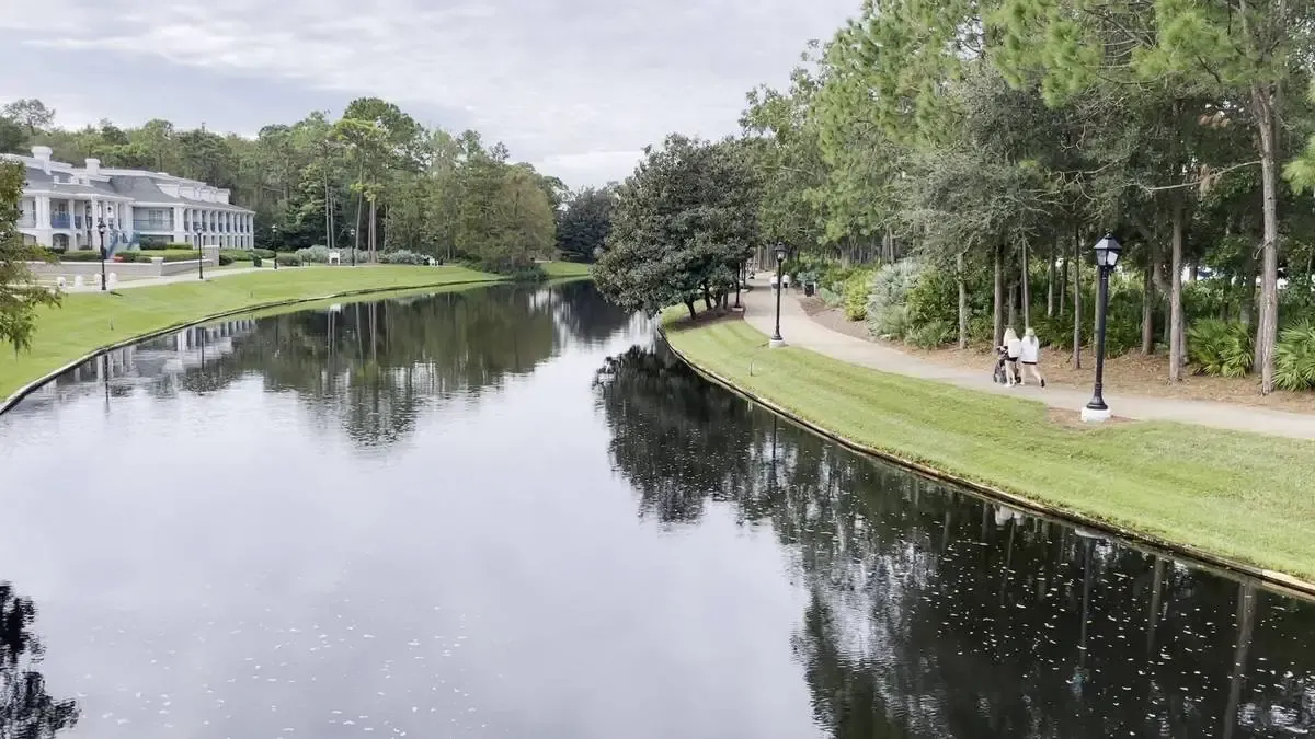 A serene canal extends through a park, bordered by a path. On the left, a large and elegant building with white columns by the water, while tall trees and lampposts line the pathway on the right.