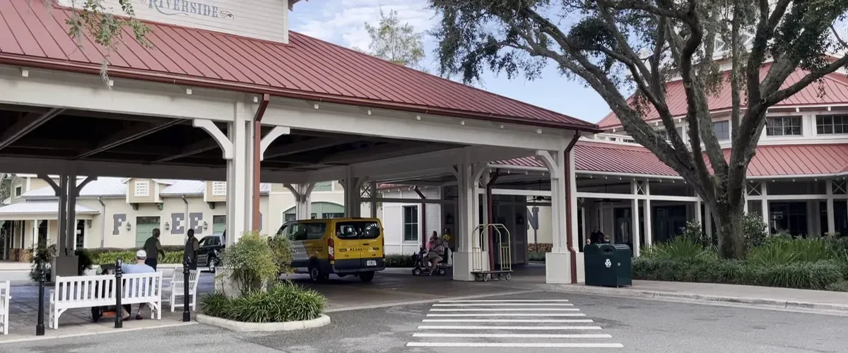 A covered entrance area with a red metal roof at a building labeled "Riverside," featuring a yellow vehicle parked nearby, people sitting on benches, and a tree providing shade.