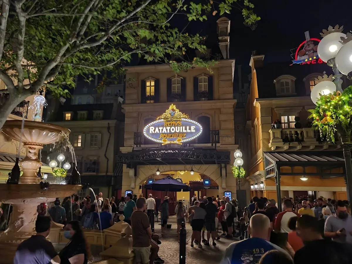A bustling nighttime scene outside Remy's Ratatouille Adventure, with a crowd gathered in front of the illuminated entrance. A decorative fountain is in the foreground, and festive lights adorn the area.
