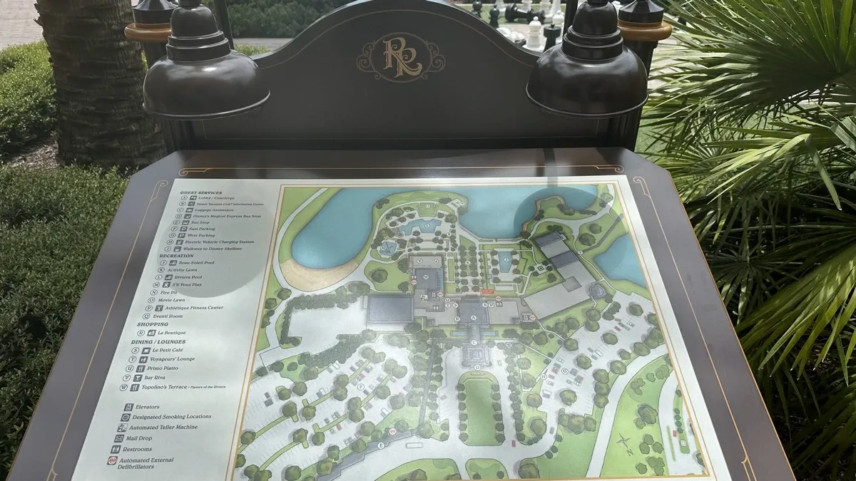A detailed outdoor map display of a resort, featuring pathways, buildings, and a pond, is illuminated by two overhead lamps. Lush greenery surrounds the map, and a large chessboard is partially visible in the background.