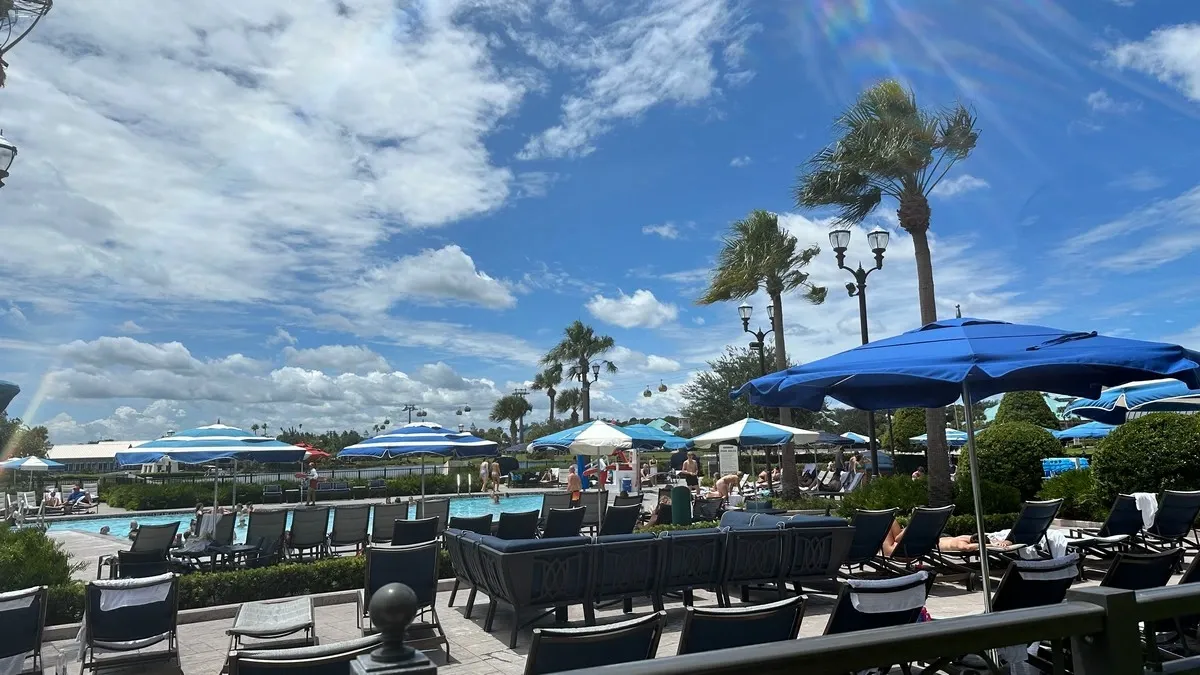 A sunny poolside scene with several blue umbrellas shading lounge chairs, surrounded by palm trees. People are relaxing and enjoying the pool under a bright blue sky with scattered clouds.