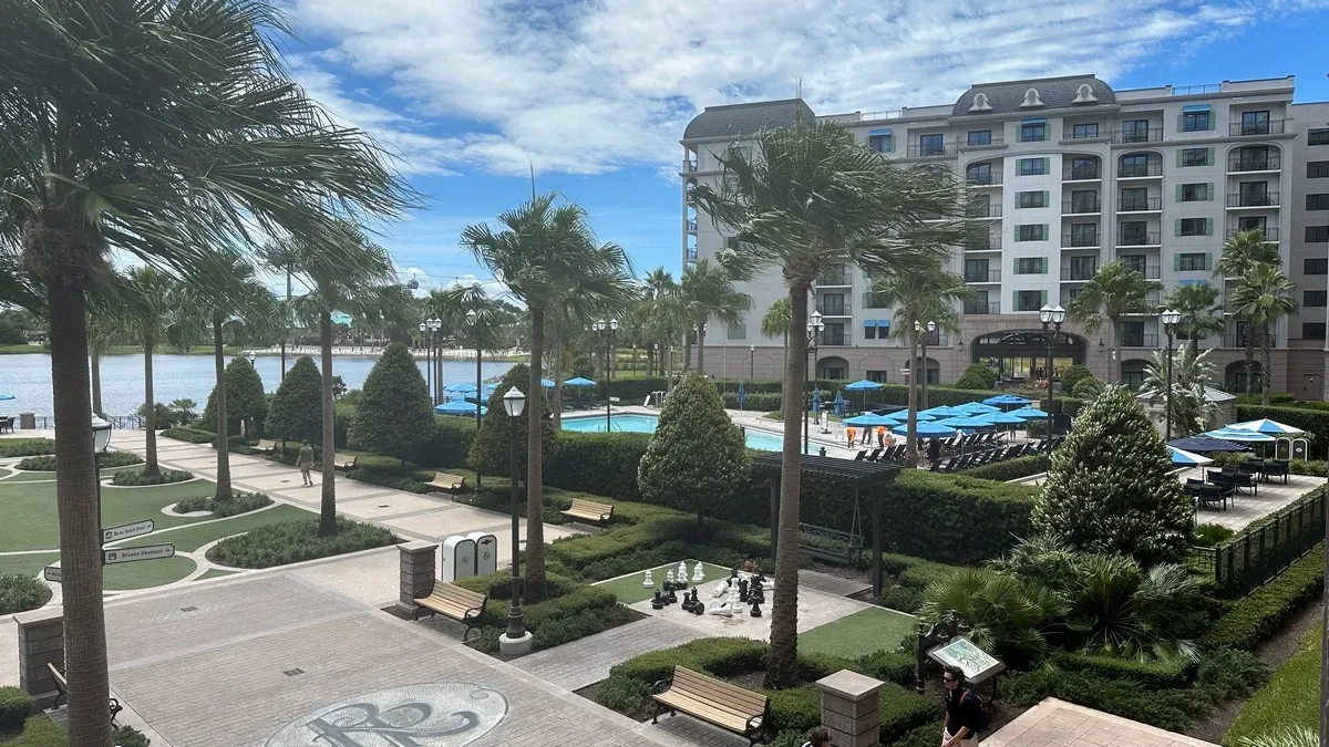 A scenic view of a resort featuring a large building with multiple floors and balconies, surrounded by lush greenery and palm trees. In the foreground, there's a paved walkway with benches and a pool can be seen through the bushes.