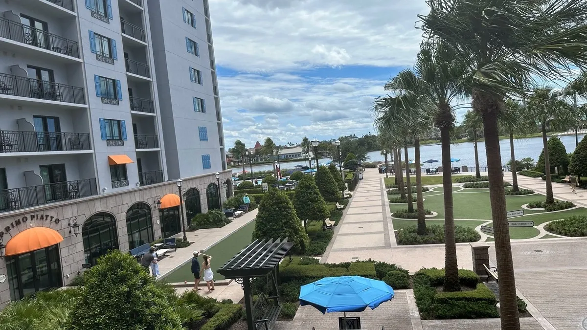 A scenic view of an hotel with a tall building featuring balconies and orange awnings. The landscaped pathway is lined with palm trees and leads to a waterfront area with blue umbrellas and neatly trimmed greenery.