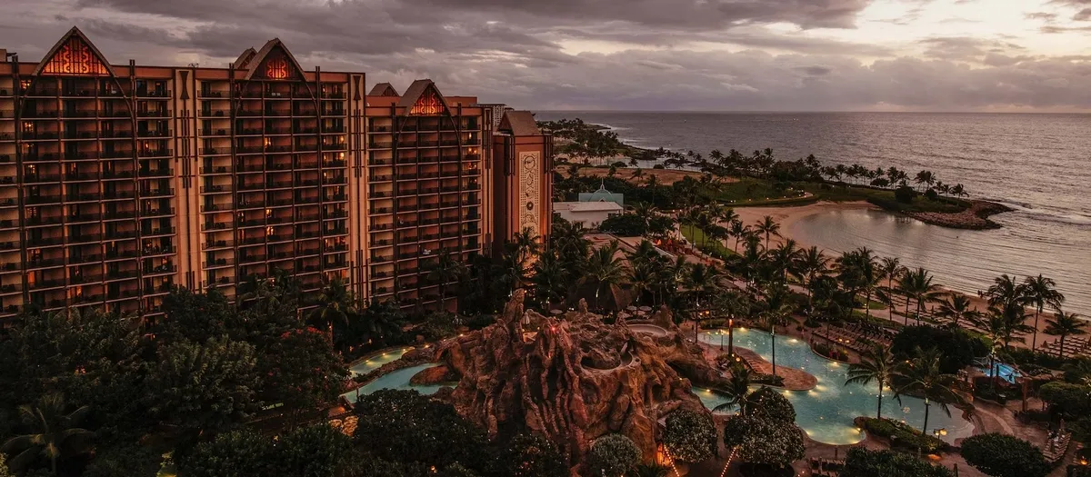 A scenic view of a large resort complex at dusk, featuring a tall building with warm lighting and a lush, tropical pool area with rock formations. The resort is set along a coastline with a calm ocean and palm trees lining the shore, under a cloudy sky.