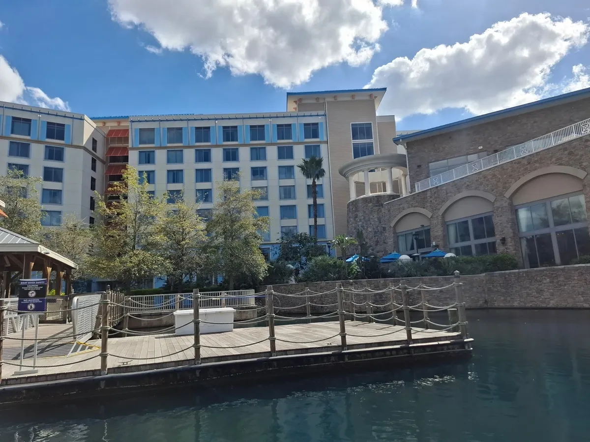 A waterfront view of an hotel with large windows, surrounded by greenery and a clear blue sky with fluffy clouds. A wooden dock with rope railings extends over the calm water, and a stone building section features arched windows and a terrace with blue umbrellas.