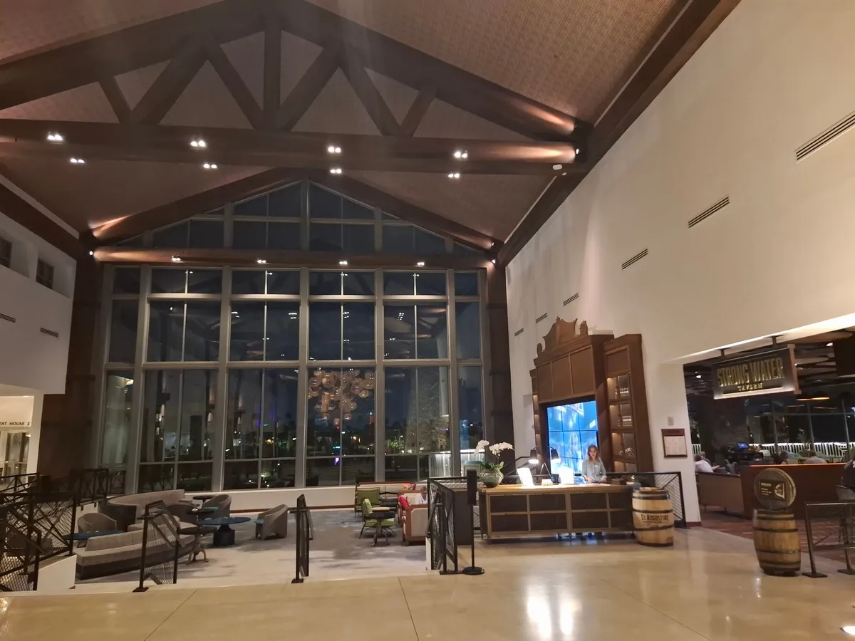 A spacious hotel lobby with a high wooden ceiling and large windows, showcasing a view of city lights at night. The area features comfortable seating and a reception desk with a staff member attending to guests. On the right, there's a restaurant entrance with a sign reading "STIRLING WHISKEY BAR & GRILL.".