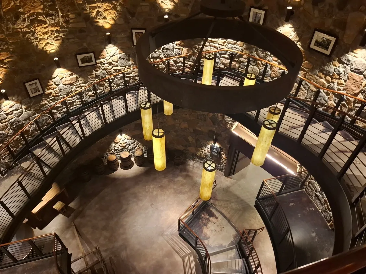 A circular room features a spiral staircase with metal railings, illuminated by large cylindrical pendant lights. The warm lighting highlights the textured walls adorned with framed pictures, and several wooden barrels are visible on the floor below.