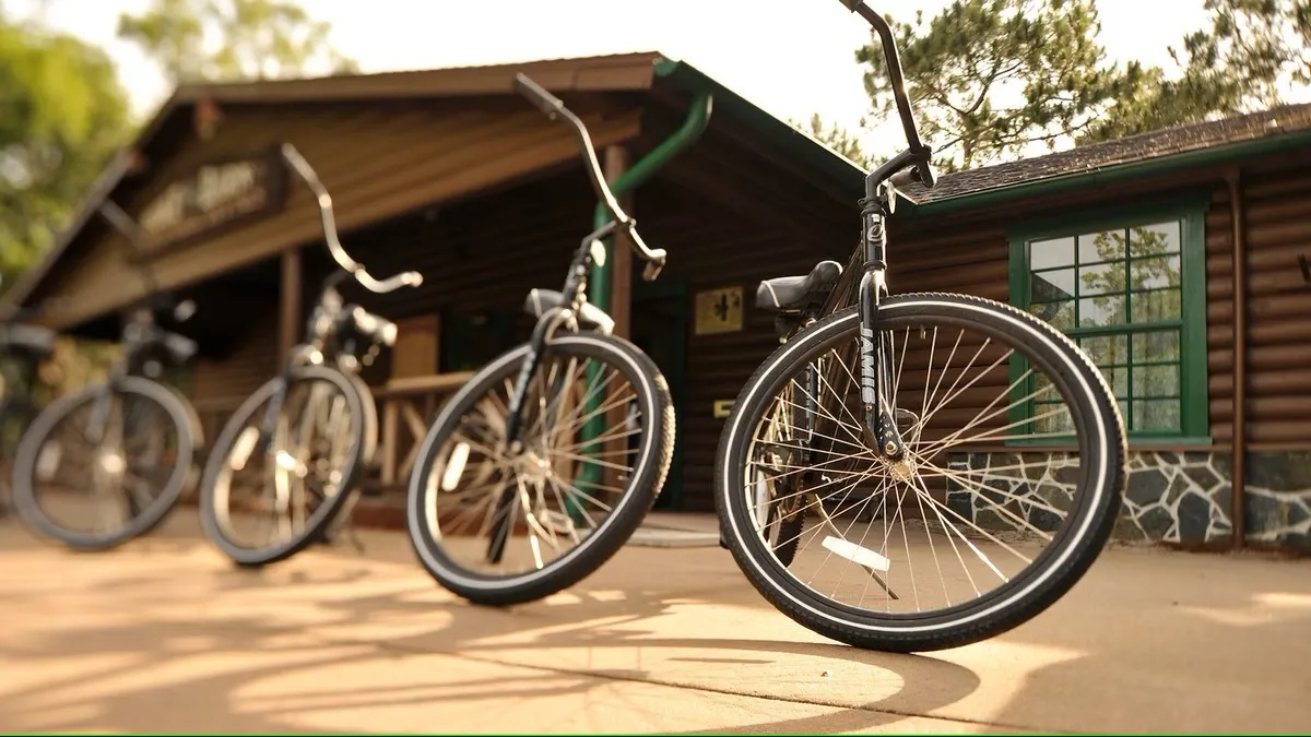Three bicycles with black frames and wide handlebars are parked in front of a rustic wooden building with green-trimmed windows, set against a backdrop of trees.