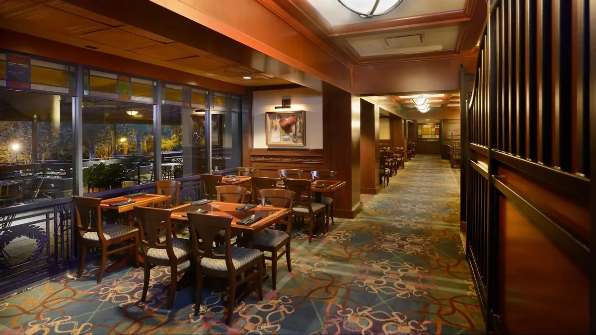A restaurant interior featuring wooden tables and chairs arranged along a patterned carpeted floor. Large windows on the left offer a view of the outside, while framed artwork adorns the walls.