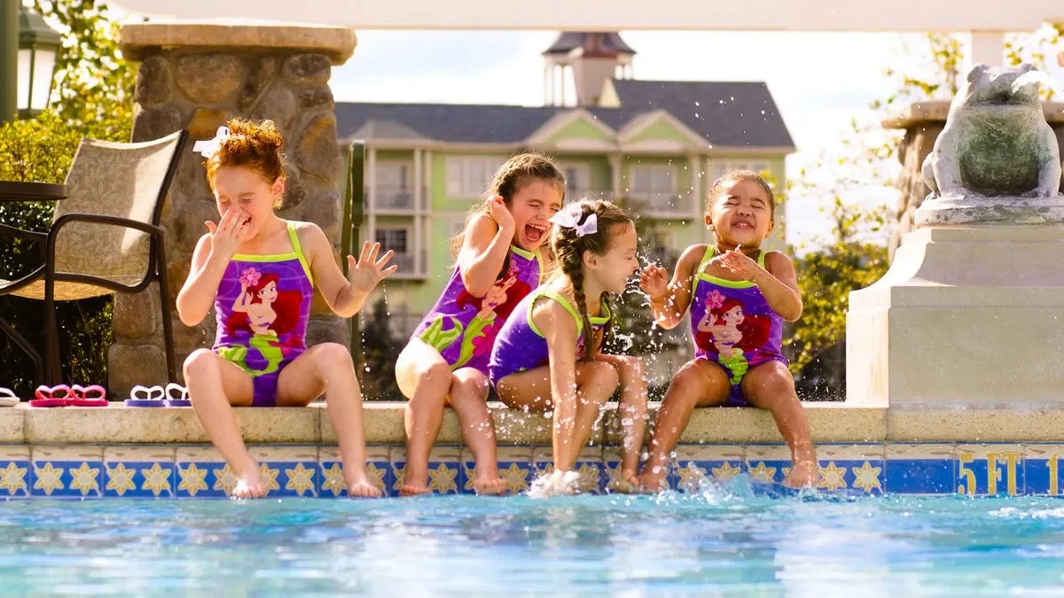 Four young girls in colorful swimsuits sit on the edge of a pool, joyfully splashing water and laughing under the bright sun, with a charming building in the background.