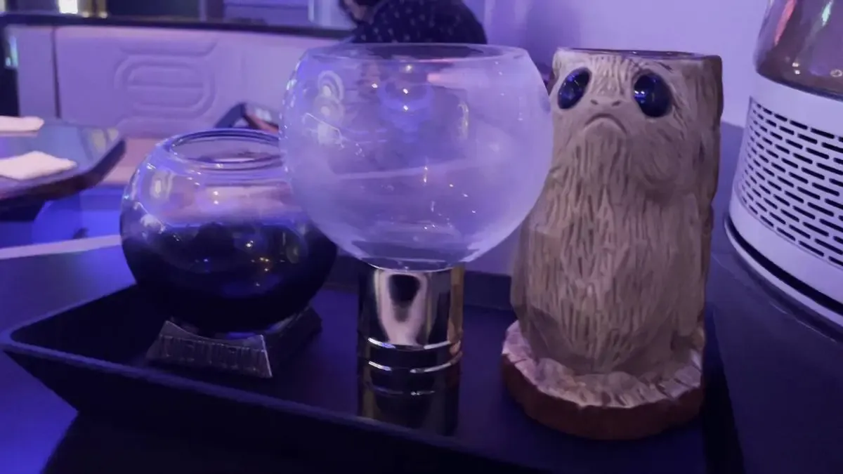 Three unique drink containers are displayed on a tray: a round glass with a dark liquid, a clear goblet with a gold base, and a wooden mug shaped like a creature with large eyes. The setting has a futuristic vibe with soft purple lighting.