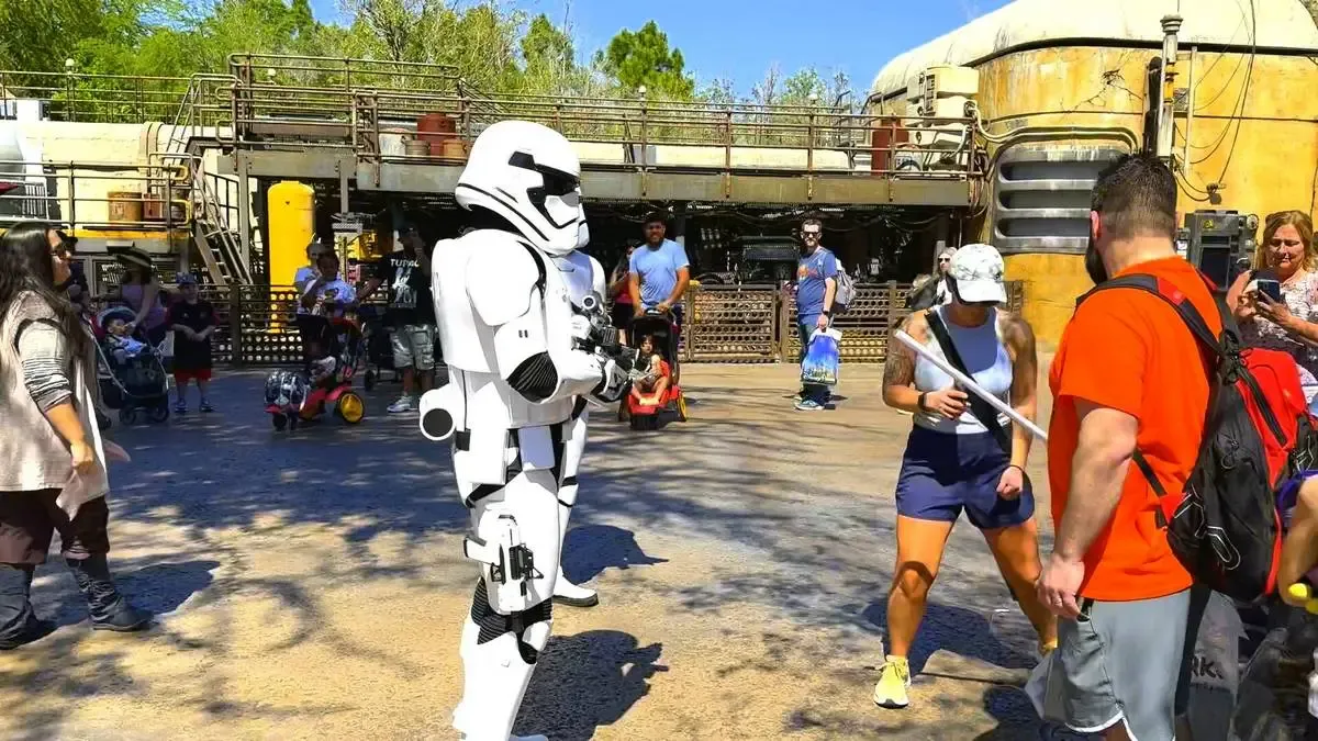 A person in a stormtrooper costume interacts with park visitors in a bustling theme park setting. The stormtrooper, dressed in iconic white armor, stands on a paved area with various people around, including a woman in athletic attire holding a lightsaber. The background features industrial-themed structures and greenery, creating an immersive atmosphere.