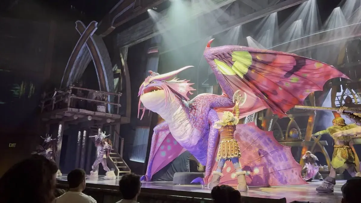 A vibrant stage performance features a large, colorful dragon with pink and purple scales and butterfly-like wings. Performers in elaborate costumes surround the dragon, set against a dramatic backdrop with theatrical lighting.
