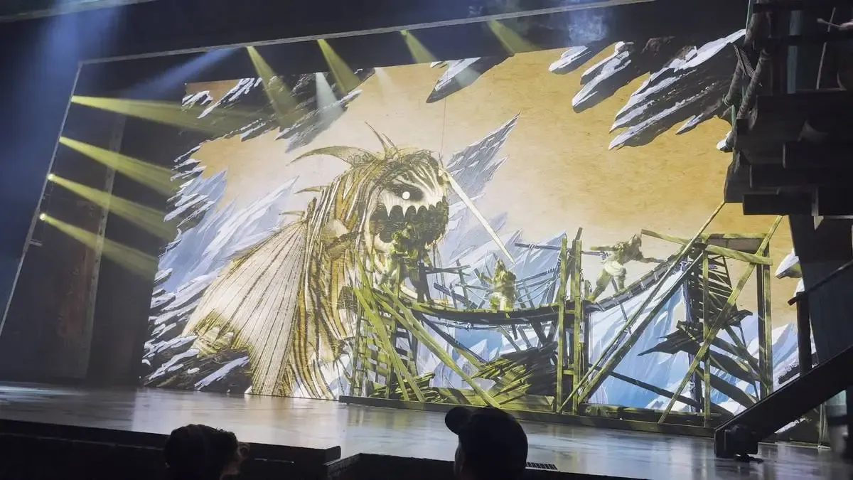 A dramatic stage scene depicts a large, menacing dragon with sharp teeth against a backdrop of jagged ice and snow. Several performers dressed in costume are positioned on a wooden, scaffold-like structure, adding to the intense atmosphere.