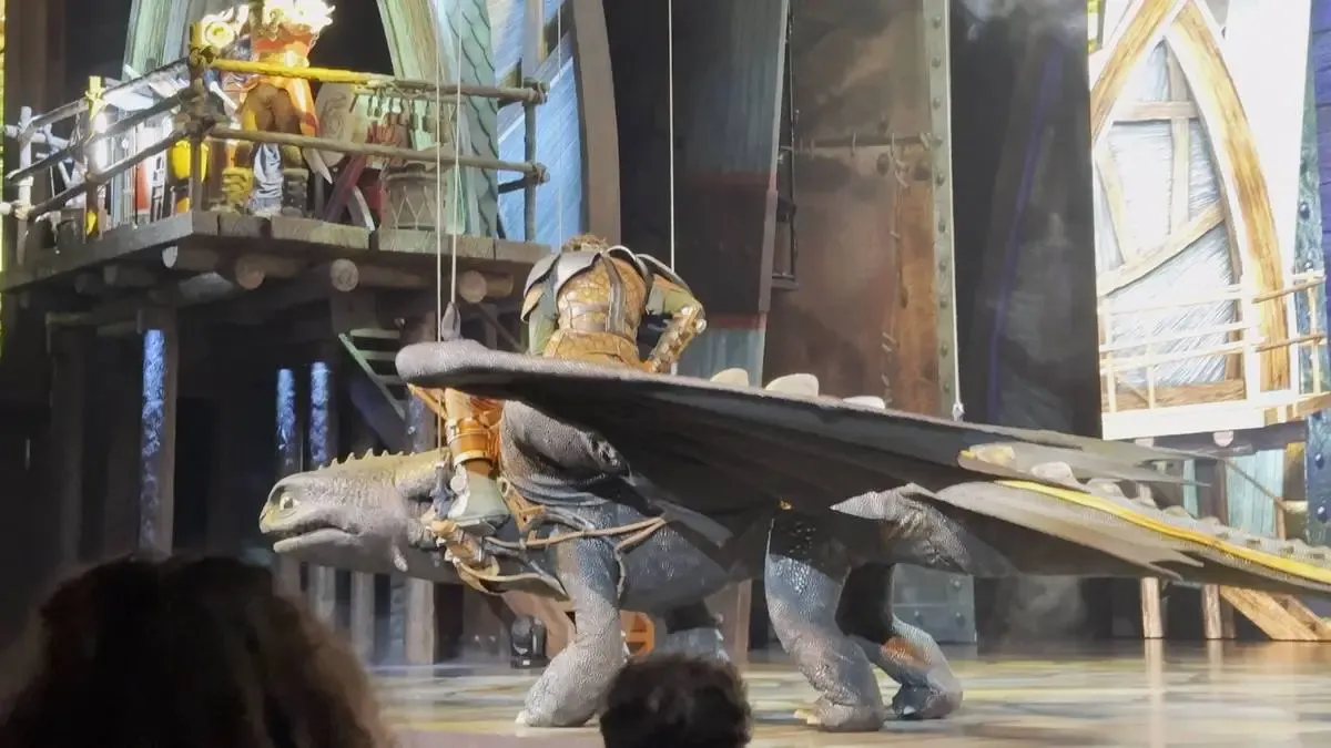 A large Toothless puppet is being ridden by a performer in a theatrical setting. The stage is designed with wooden structures and a balcony where another character stands.