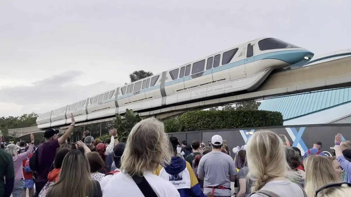 A crowd of people looks up and waves at a sleek, white monorail with a blue stripe as it glides along an elevated track. The scene captures a moment of excitement and interaction, with a cloudy sky in the background.