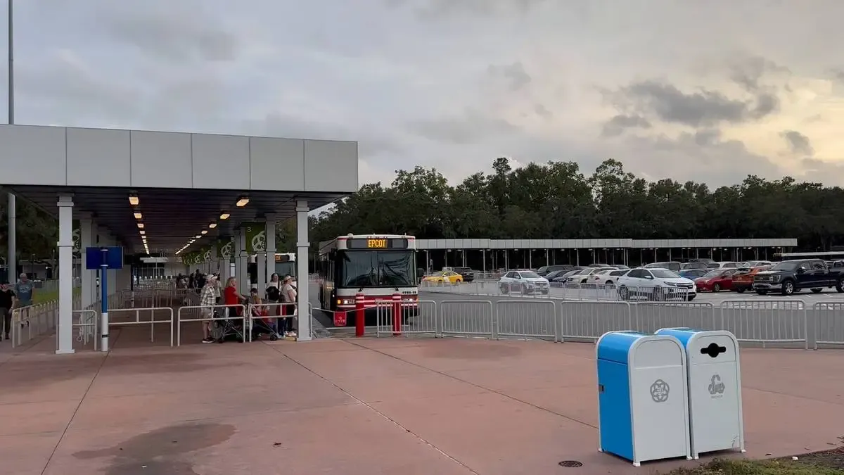 A bus labeled "Epcot" is parked at a station with a covered waiting area where people are seated. The setting is a parking lot with several cars, and the sky is overcast. Nearby, there are two trash bins, one blue and one white.