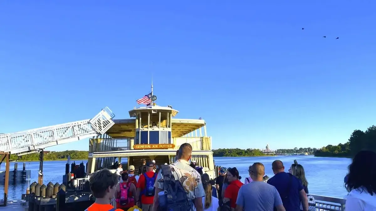 A group of people boards the ferryboat "General Joe Potter" on a clear day, with a calm blue lake and distant trees in the background. An American flag waves atop the boat, and a few birds fly in the bright blue sky.
