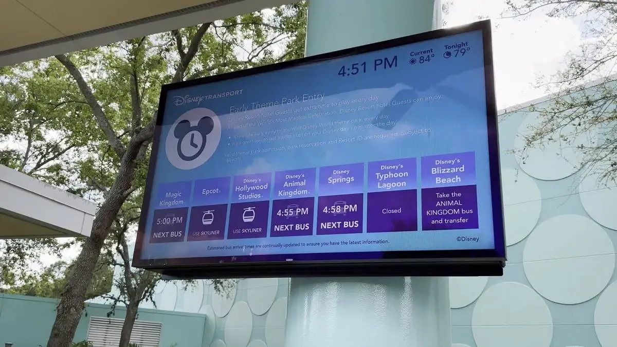 A digital display at a Disney Transport station shows schedules for various destinations like Magic Kingdom, Epcot, and Disney Springs, with specific departure times and notes about transportation options. The screen is mounted on a pole, with trees and part of the station's architecture visible in the background.
