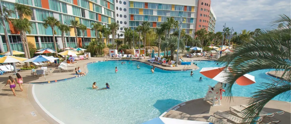 A resort pool area featuring people enjoying the water, surrounded by colorful sun umbrellas and palm trees. The backdrop includes a modern building with a colorful, geometric facade, creating a lively and inviting atmosphere.