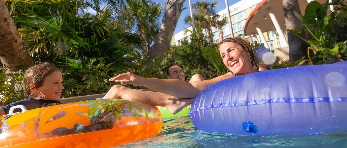 A smiling woman and a child are enjoying a pool, each floating on colorful inflatable tubes, surrounded by lush greenery and a building in the background.
