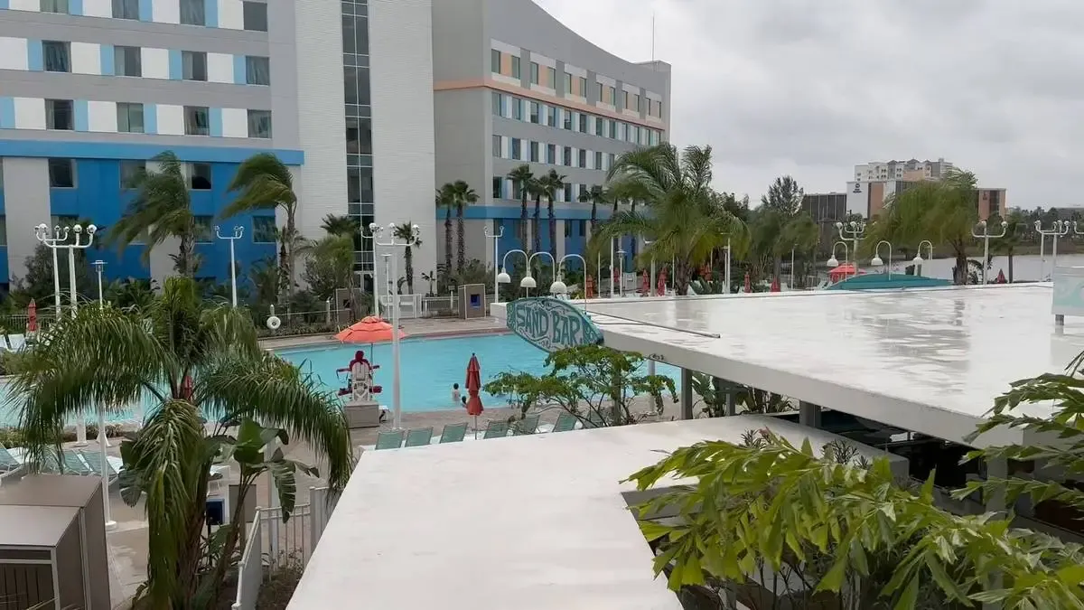 A modern hotel with a large swimming pool surrounded by palm trees and lounge chairs. The building features a mix of gray, blue, and white panels, and the area appears calm under a cloudy sky. A sign for a "Sand Bar" is visible near the pool.