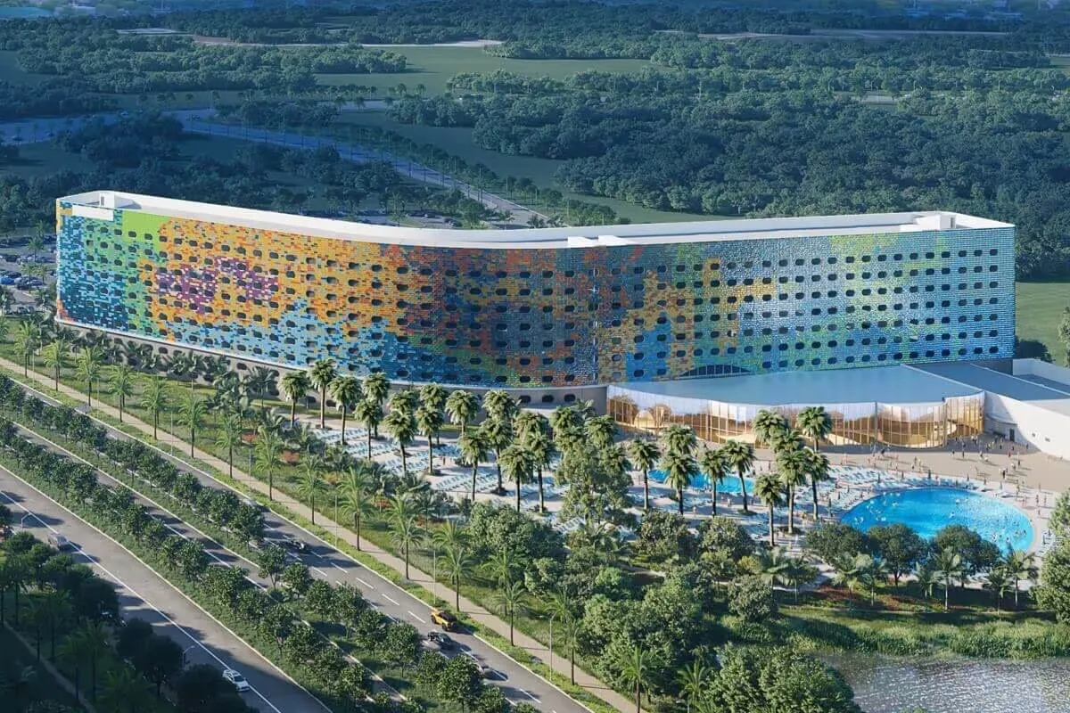 Aerial view of a vibrant, multi-colored hotel with a unique, perforated facade. In front, a row of palm trees lines a large swimming pool area, while a multi-lane road runs alongside the property.