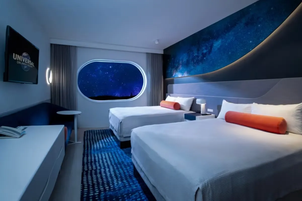 A modern hotel room features two neatly made beds with orange bolster pillows, set against a space-themed wall mural. A flat-screen TV and a small table with chairs complete the stylish decor.