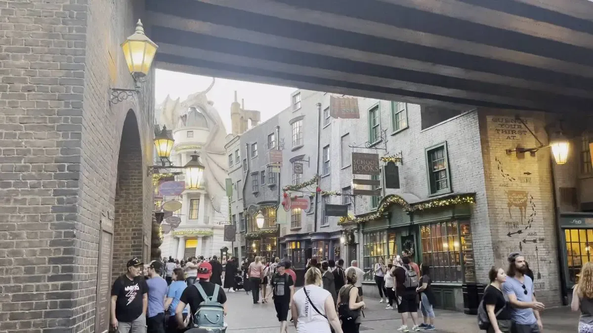 A street scene at a themed attraction, featuring a cobblestone alley lined with quaint shops. A large dragon sculpture looms above a distant building, adding a whimsical touch.