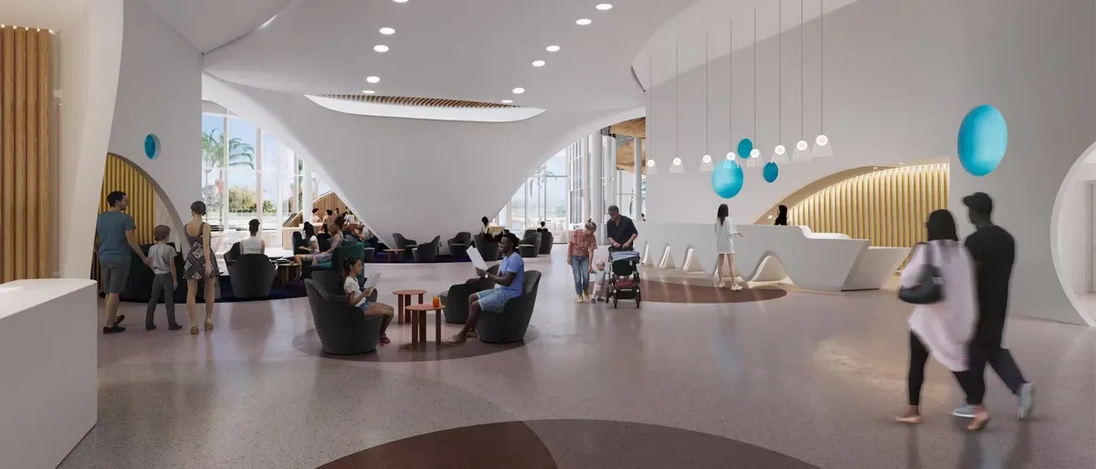 A modern, spacious lobby features people walking and sitting on sleek furniture. The room has a high ceiling with circular light fixtures, a curved white reception desk, and blue decorative accents. Large windows let in natural light, and a few people are gathered near the entrance.