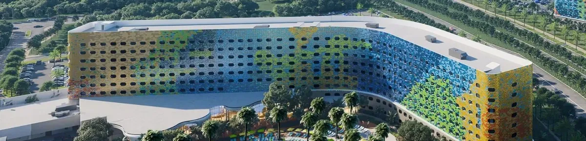 Aerial view of a large, curved building with a vibrant, multicolored facade featuring shades of blue, green, and yellow. The structure is surrounded by lush greenery and palm trees, with a parking lot visible in the background.