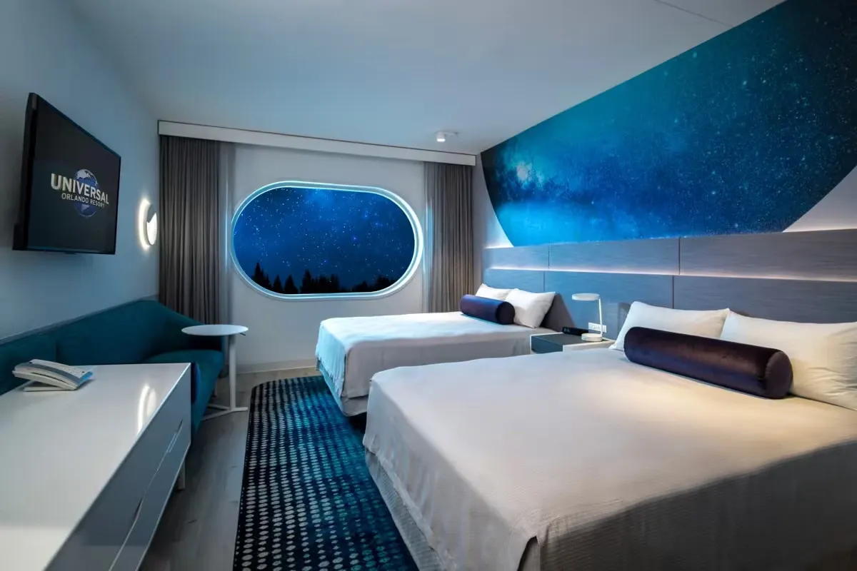 A modern hotel room features two beds with purple bolster pillows and a striking space-themed mural on the wall. A large, oval window displays a starry night sky, enhancing the room's cosmic theme. A flat-screen TV is mounted on the wall, and a small seating area with a teal sofa and round table adds to the cozy ambiance.