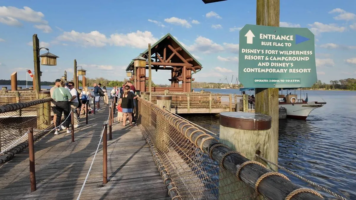People walking along a wooden dock with rustic railings, leading to a boat landing. A sign indicates directions to Disney's Fort Wilderness Resort & Campground and Disney's Contemporary Resort.