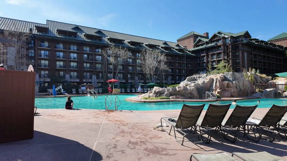 An outdoor pool area with lounge chairs is set against a large, rustic hotel with green roofs. The pool is surrounded by natural rock formations, and a few people are enjoying the water under a clear blue sky.