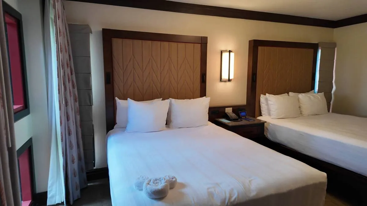 A cozy hotel room featuring two beds with white linens and multiple pillows, separated by a wooden nightstand with a phone. A soft light fixture is mounted on the wall, and the room is accented with patterned curtains and a warm, neutral color palette.