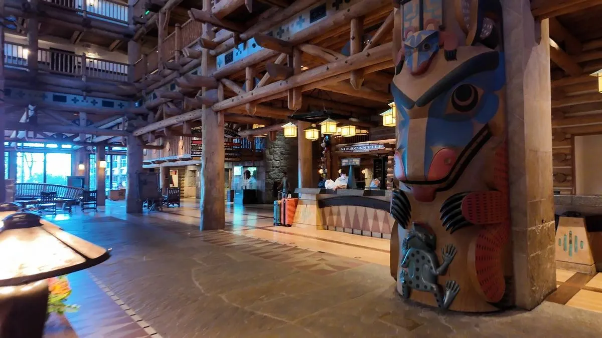 A spacious lodge interior featuring rustic wooden beams and a colorful totem with intricate designs. Large windows and wooden furniture complete the cozy atmosphere.