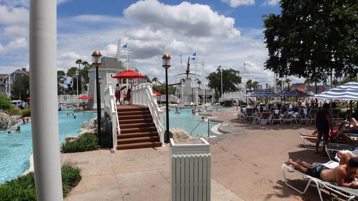 A pool area featuring a bridge with red umbrellas and lifeguards, surrounded by people swimming and lounging on chairs.