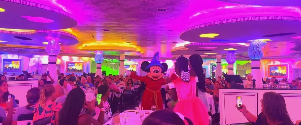 A lively dining scene features a costumed character in a red robe entertaining guests in a colorful, illuminated restaurant. The room is filled with people, some capturing the moment with their phones, while vibrant pink and purple lights create a festive atmosphere.
