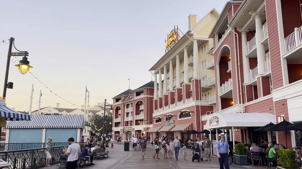 People strolling along a boardwalk lined with red and white buildings featuring balconies and columns, as evening sets in with soft lighting from street lamps.