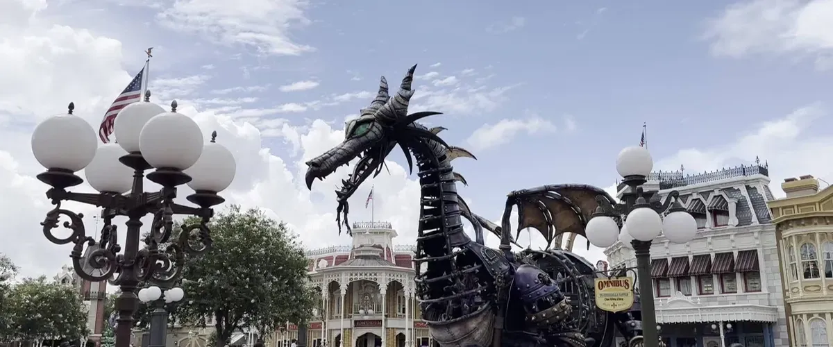 A large dragon float with metallic scales and wings is part of a parade in a theme park, surrounded by vintage-style street lamps and colorful, ornate buildings under a partly cloudy sky.