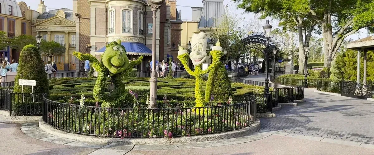 Topiary sculptures of Lumiere and Din Don stand cheerfully in a garden, surrounded by a black wrought-iron fence. The setting is a theme park area with colorful buildings and a few visitors strolling by.
