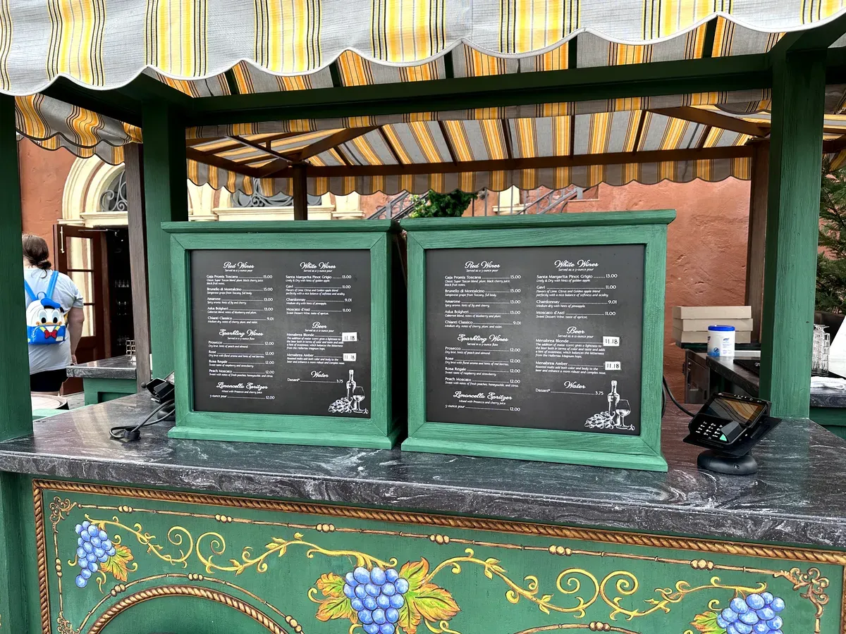 A charming outdoor kiosk with a striped canopy displays two green-framed menus listing various wines and beers. The counter is adorned with painted grapevine designs, and a payment terminal is visible on the right.
